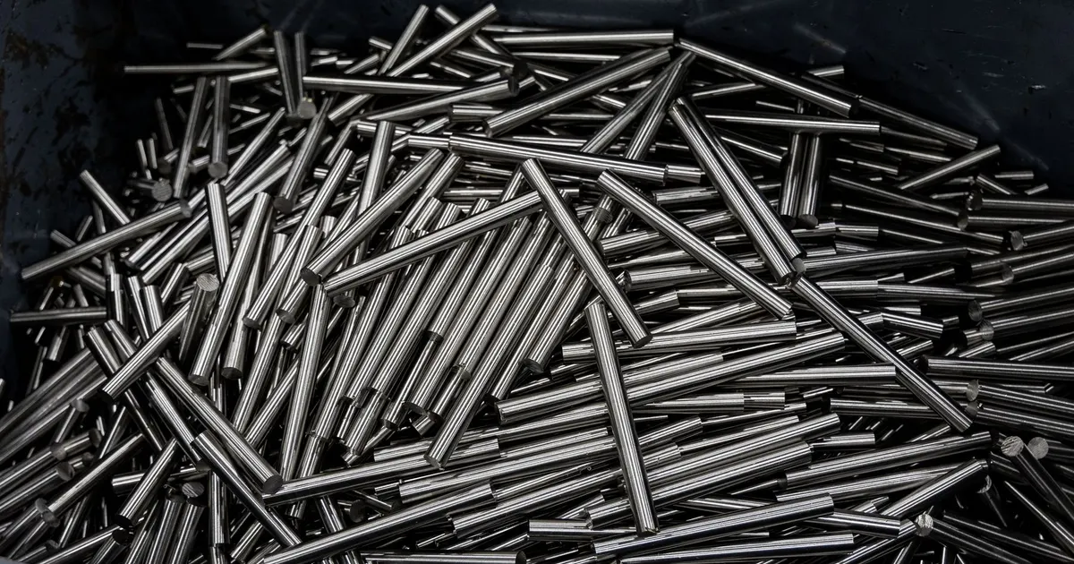 Pile of bright metal rods in a dark bin, the kind of bar stock cut into parts that later need passivation