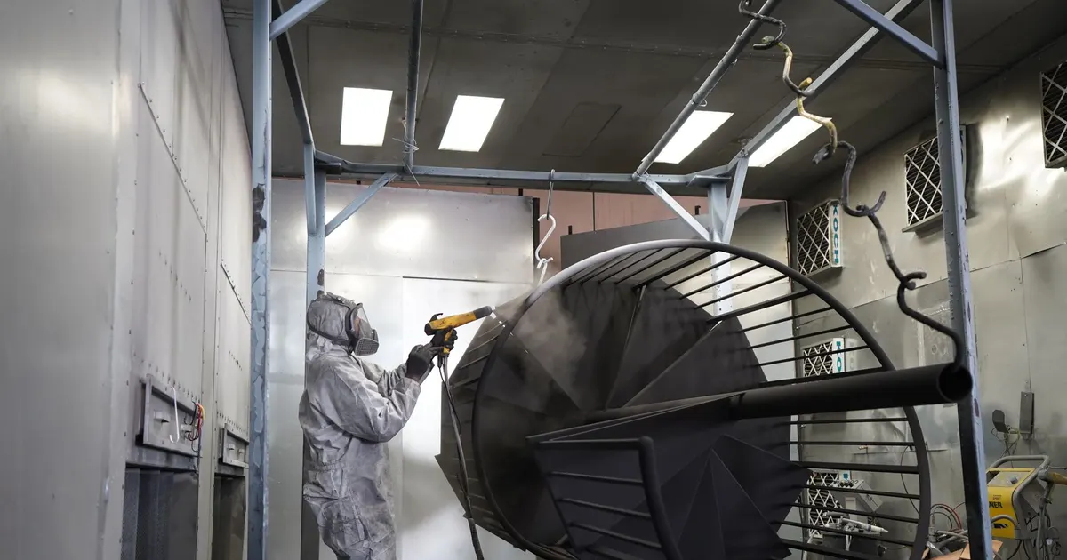 Technician in a protective suit and respirator spraying coating onto a black metal frame in a paint booth